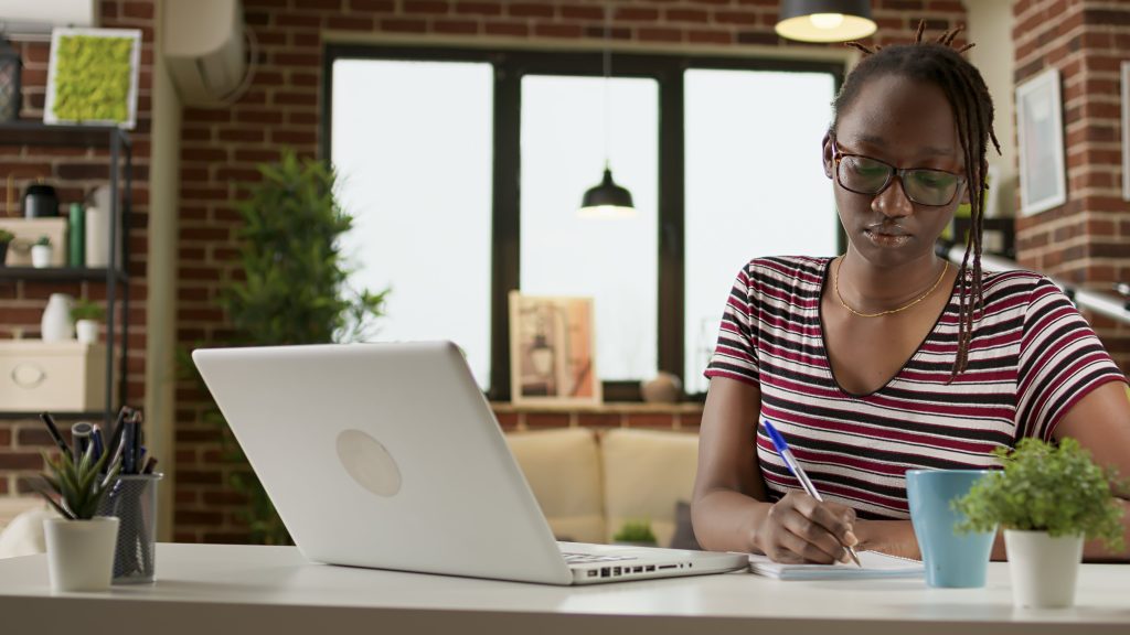 Mulher jovem, negra que escreve em um papel pensativa enquanto está com um computador aberto em sua mesa representando os estudos para Residência Médica em 2023. 
