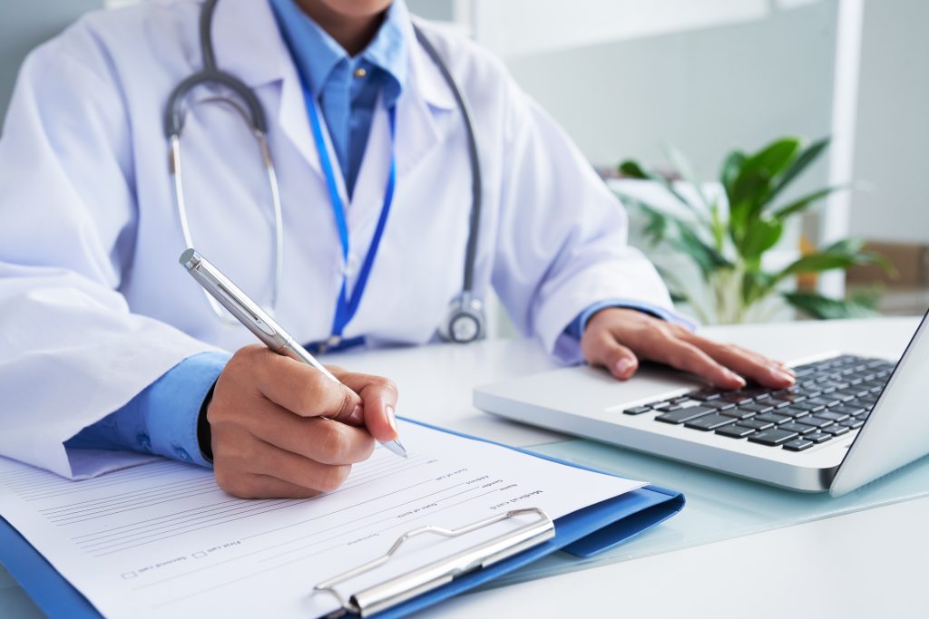 Médico com jaleco sentado em frente a um computador enquanto escrev algo em um papel. Disciplina no estudo para a Residência Médica. 