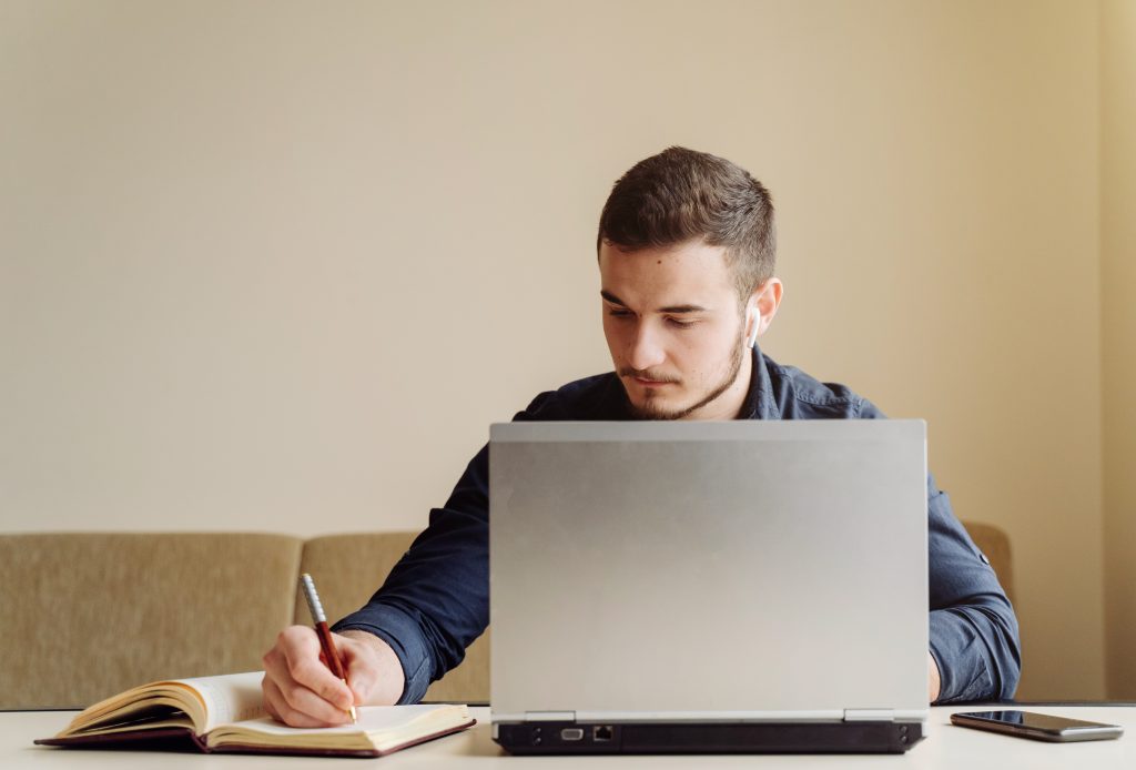 Rapaz jovem sentado em frente ao computador enquanto escreve algo em sua agenda e lê em seu computador. Foto ilustrando como corrigir os erros da sua prova de Residência Médica. 