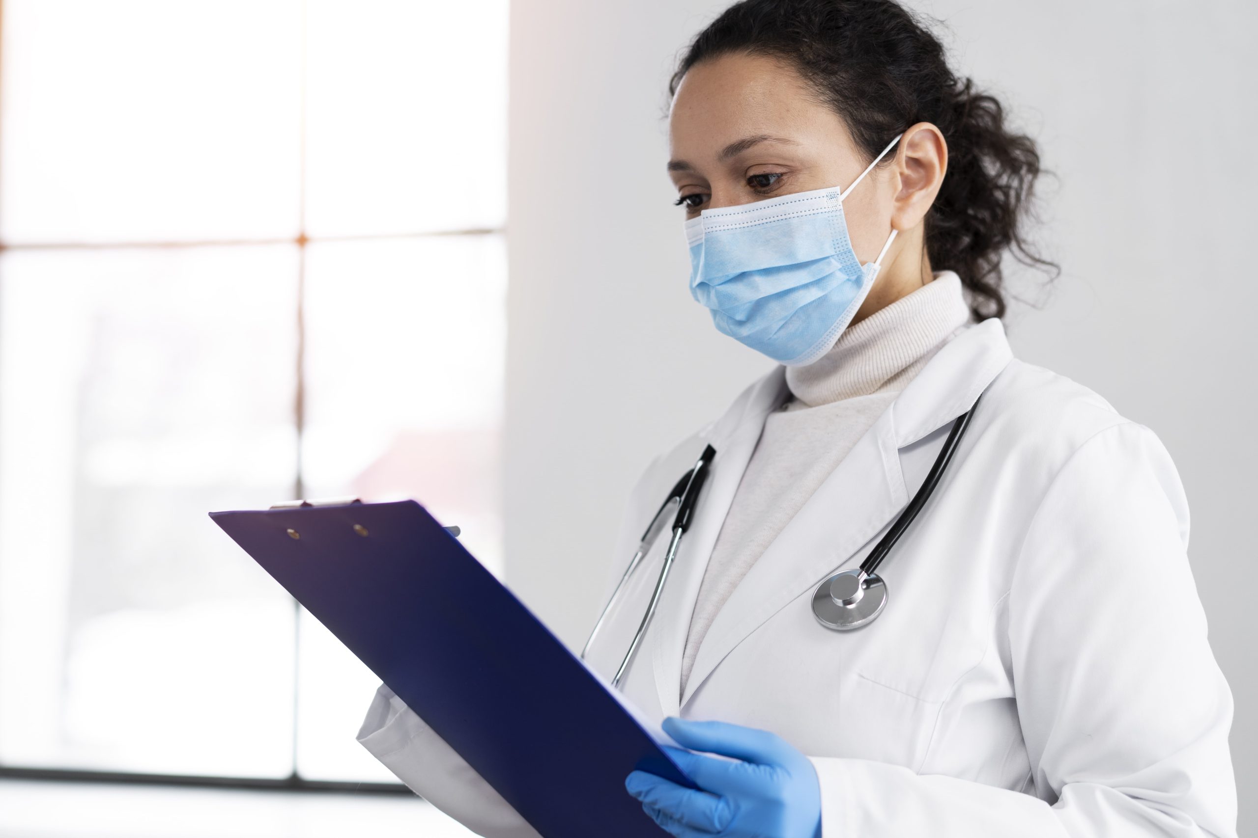 Médica com uma prancheta na mão escrevendo vestida de branco e portando um estetoscópio no pescoço (Provas de Residência Médica de SP)